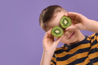 Photo of Boy covering eyes with halves of fresh kiwi on violet background, space for text