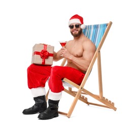 Muscular young man in Santa hat with deck chair, gift box, sunglasses and cocktail on white background