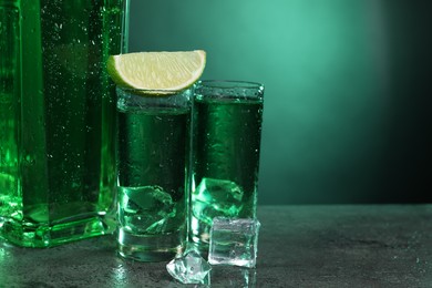 Photo of Absinthe in shot glasses, lime and ice cubes on gray textured table against green background, closeup with space for text. Alcoholic drink