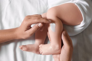 Photo of Mother applying moisturizing cream onto baby`s leg on bed, top view