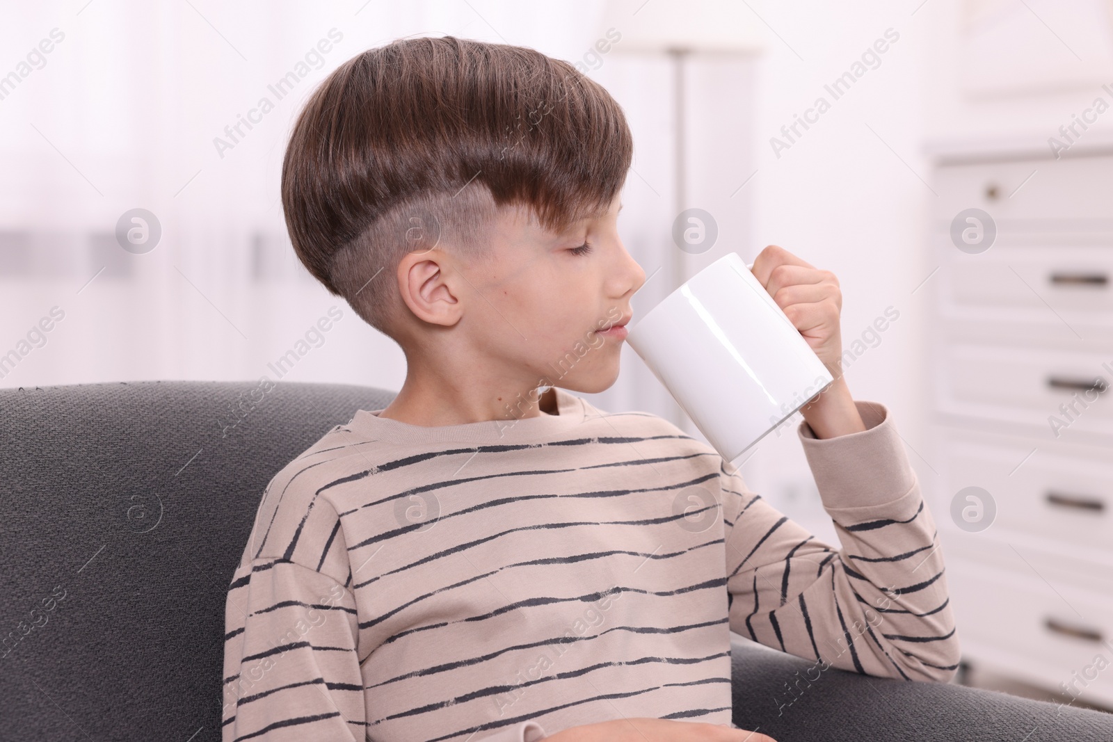 Photo of Cute boy drinking from white ceramic mug on sofa at home