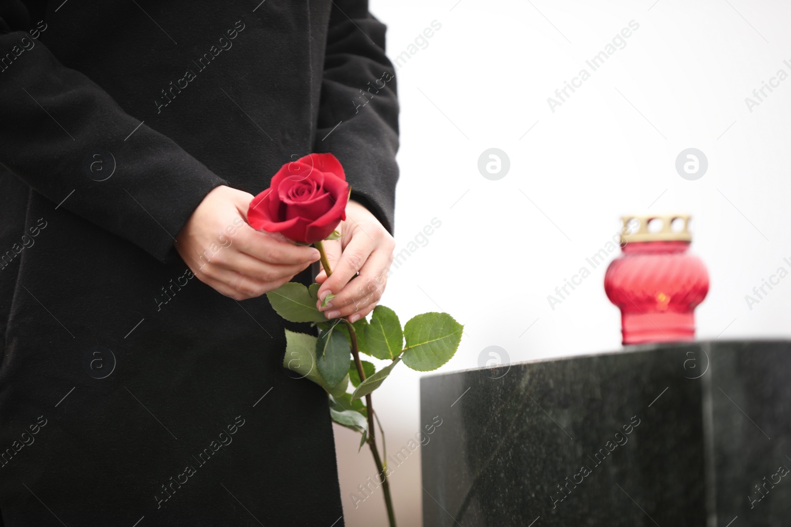 Photo of Woman holding red rose near black granite tombstone with candle outdoors, closeup. Funeral ceremony