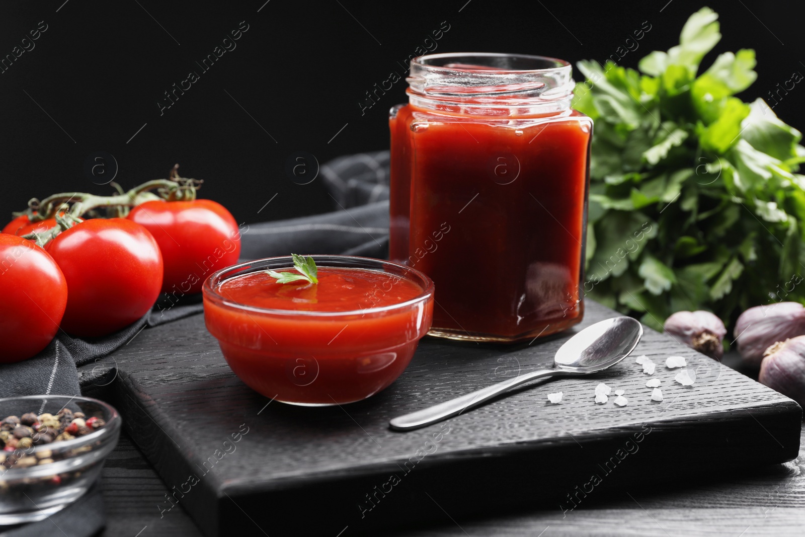 Photo of Delicious ketchup, spoon and spices on black wooden table. Tomato sauce