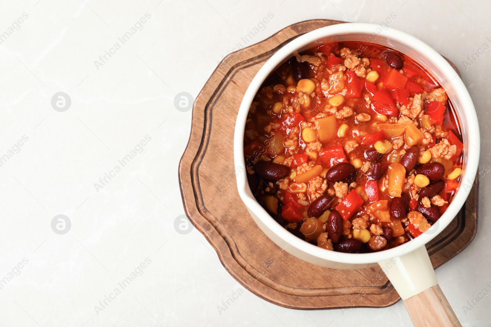 Photo of Saucepan with chili con carne, wooden board and space for text on light background, top view