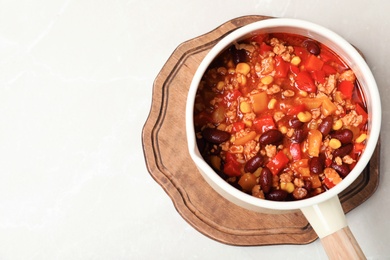 Photo of Saucepan with chili con carne, wooden board and space for text on light background, top view