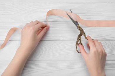 Photo of Woman cutting beige ribbon with scissors at white wooden table, closeup