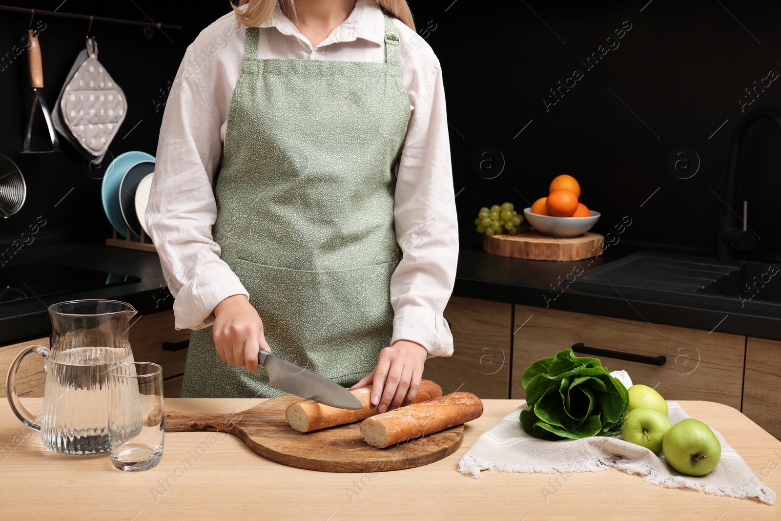 Photo of Woman in clean apron cutting baguette on wooden table at kitchen, closeup