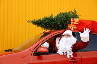 Authentic Santa Claus with fir tree and bag full of presents driving car against yellow background