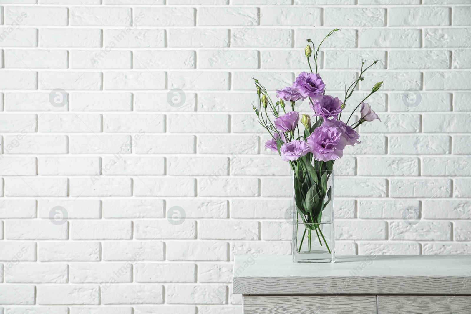 Photo of Vase with beautiful Eustoma flowers on table against brick wall