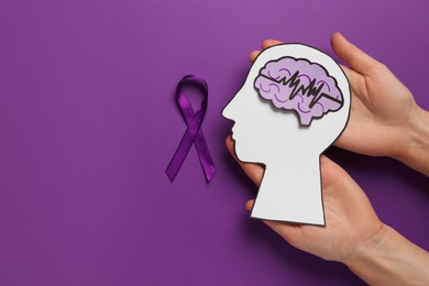 Photo of Top view of woman holding paper human head cutout with brain near purple ribbon on color background, space for text. Epilepsy awareness