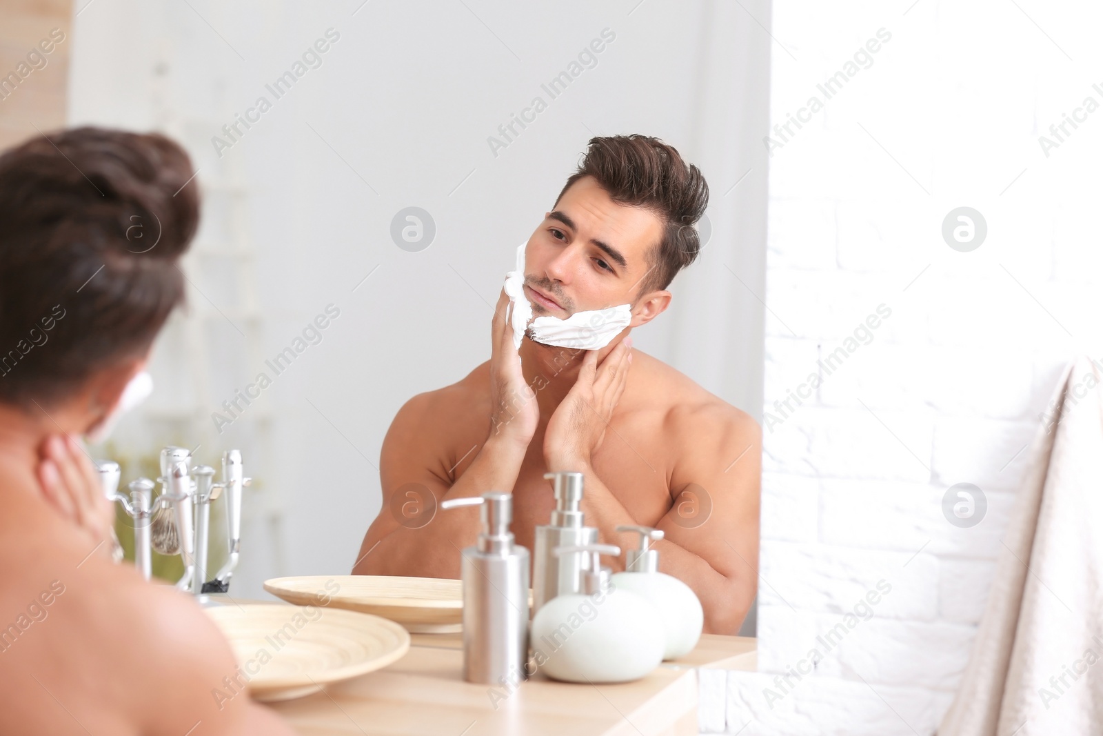 Photo of Young man applying shaving foam near mirror in bathroom