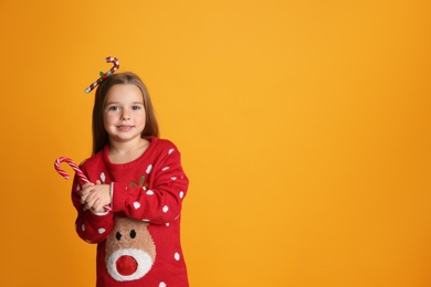Cute little girl in Christmas sweater and festive headband holding candy cane on yellow background, space for text