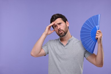 Photo of Unhappy man with hand fan suffering from heat on purple background. Space for text