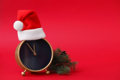 Alarm clock in Santa hat and fir branch on red background, space for text. New Year countdown