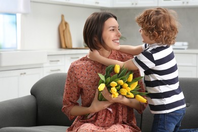 Little son congratulating his mom with Mother`s day at home, space for text. Woman holding bouquet of yellow tulips