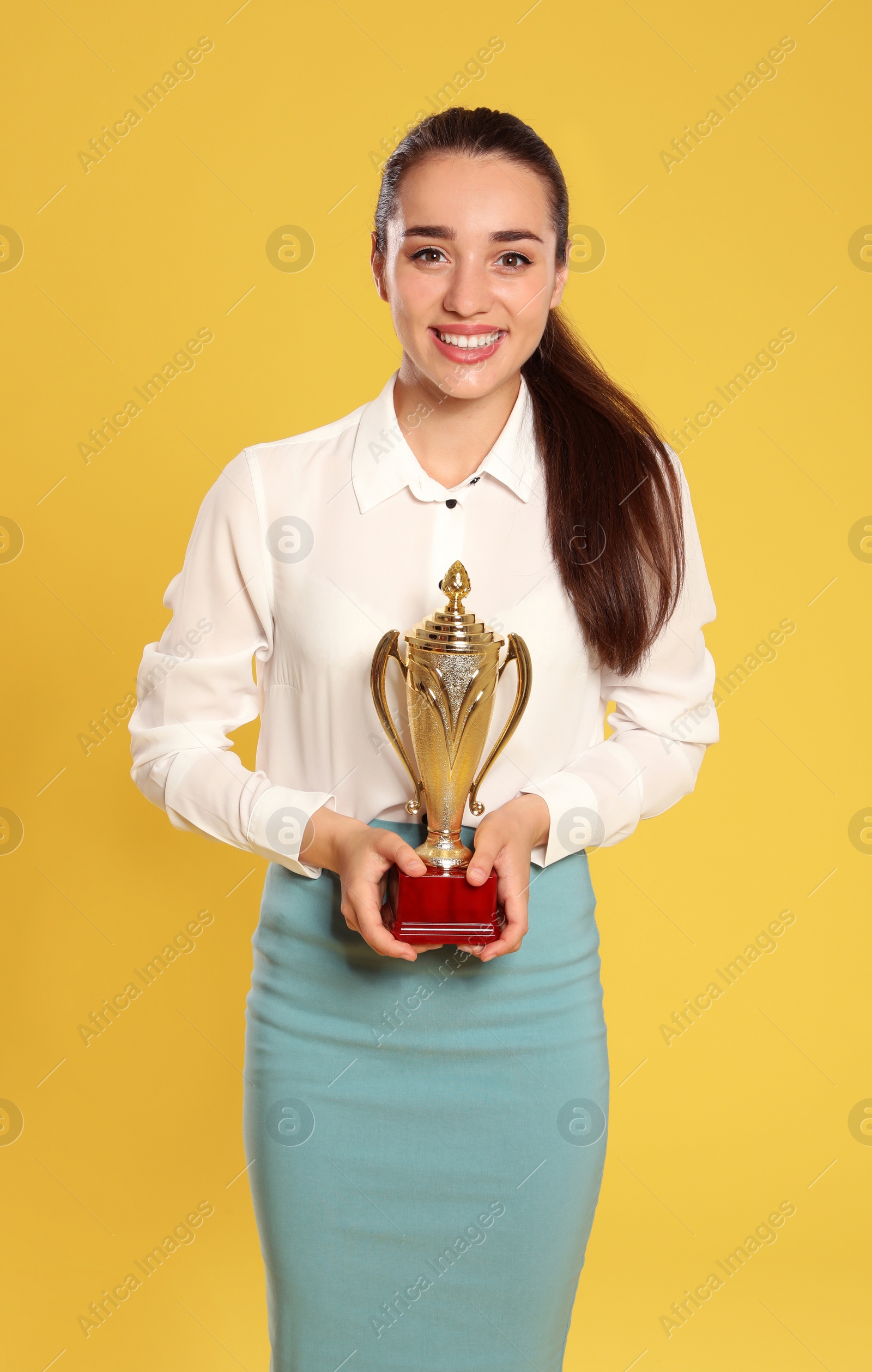 Photo of Portrait of happy young businesswoman with gold trophy cup on yellow background