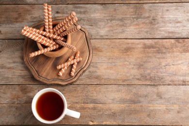 Photo of Chocolate wafer rolls in bowl and cup of tea on wooden table, top view with space for text