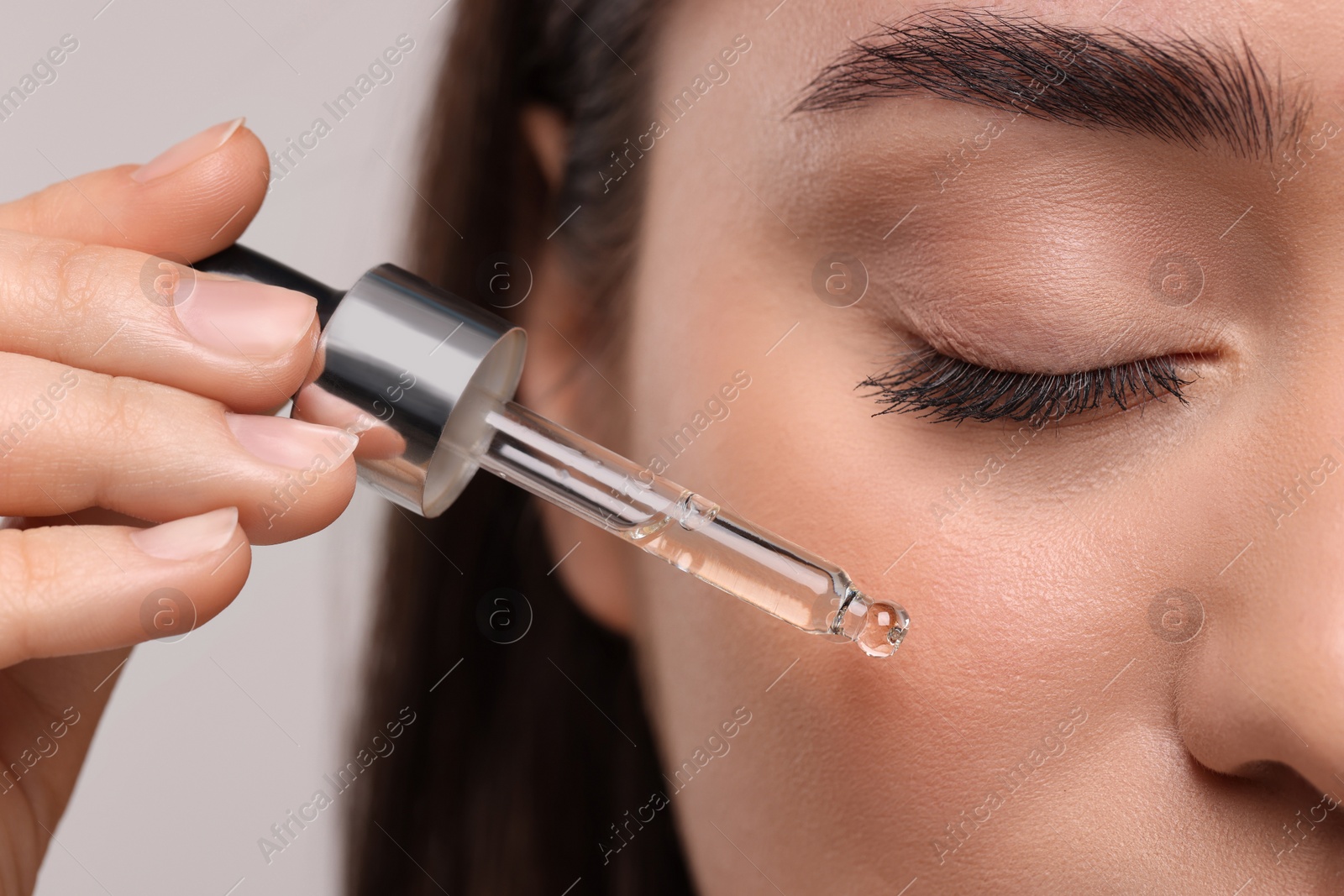 Photo of Young woman applying essential oil onto face on light grey background, closeup