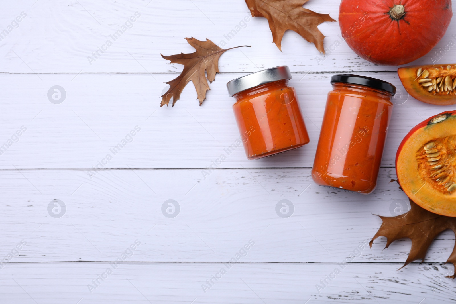 Photo of Jars of delicious pumpkin jam, fresh pumpkins and dry leaves on white wooden table, flat lay. Space for text