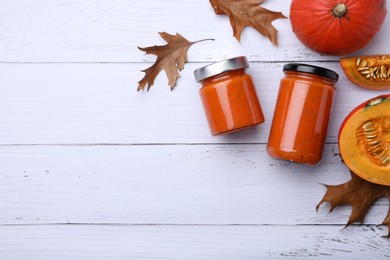 Photo of Jars of delicious pumpkin jam, fresh pumpkins and dry leaves on white wooden table, flat lay. Space for text