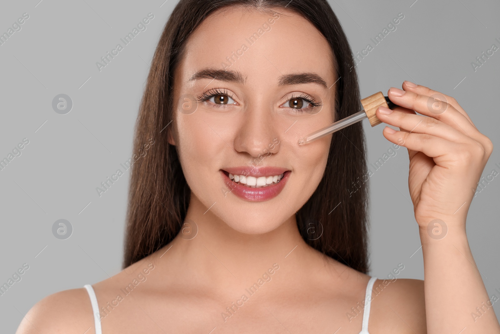 Photo of Beautiful young woman applying essential oil onto face on grey background