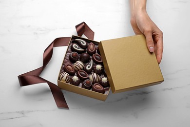 Photo of Woman with open box of delicious chocolate candies at white marble table, top view