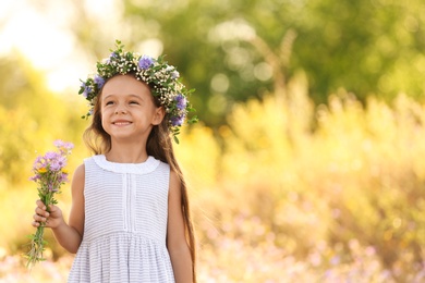 Photo of Cute little girl wearing beautiful wreath with bouquet of wildflowers outdoors, space for text. Child spending time in nature