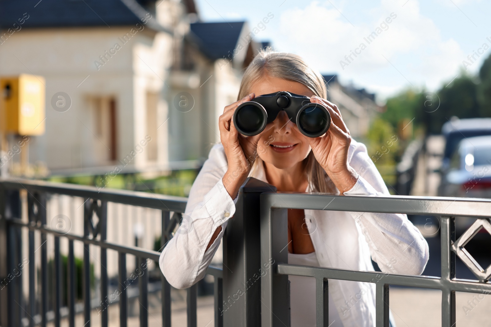 Photo of Concept of private life. Curious senior woman with binoculars spying on neighbours over fence outdoors