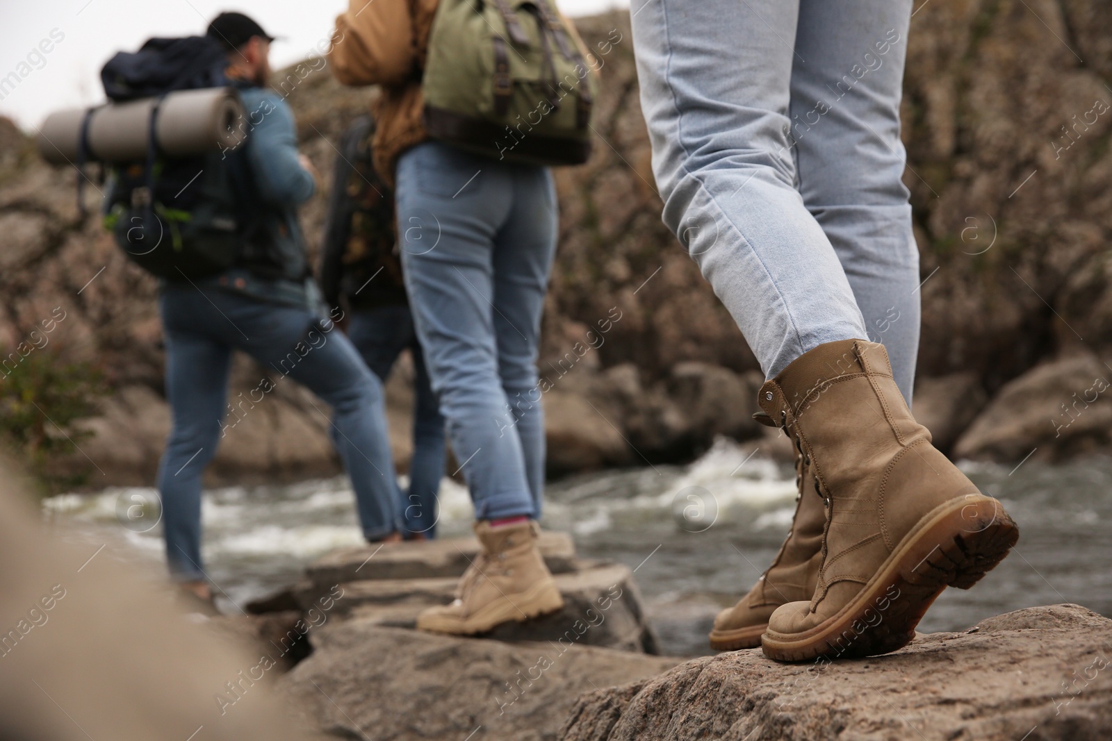 Photo of Group of friends with backpacks crossing mountain river, focus on hiking boots