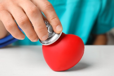 Doctor holding stethoscope near red heart on wooden table, closeup. Cardiology concept