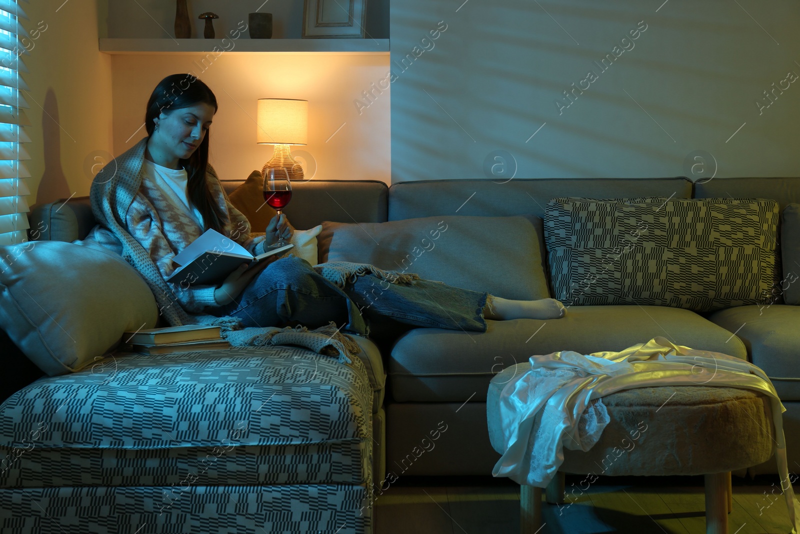 Photo of Woman with glass of wine reading book on couch in room at night