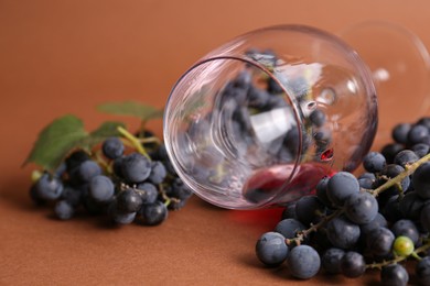 Photo of Overturned glass with red wine and grapes on brown background, closeup
