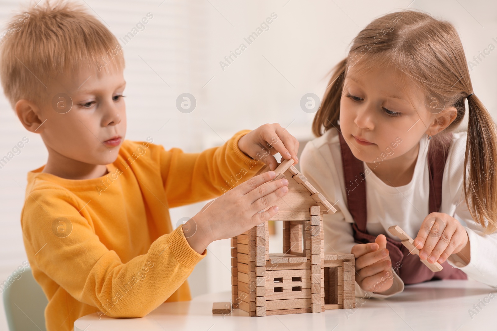 Photo of Little boy and girl playing with wooden house at white table indoors. Children's toys