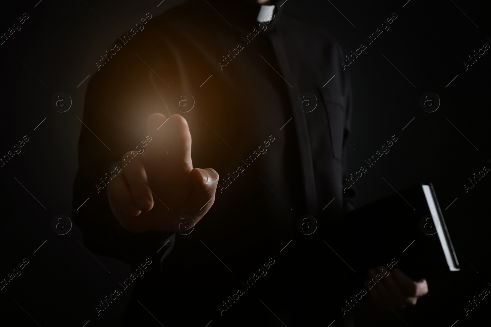 Image of Priest with Bible making blessing gesture and holy light on dark background, closeup