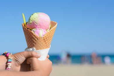 Photo of Little girl holding waffle cone with scoops of delicious colorful ice cream at beach on sunny summer day, closeup. Space for text