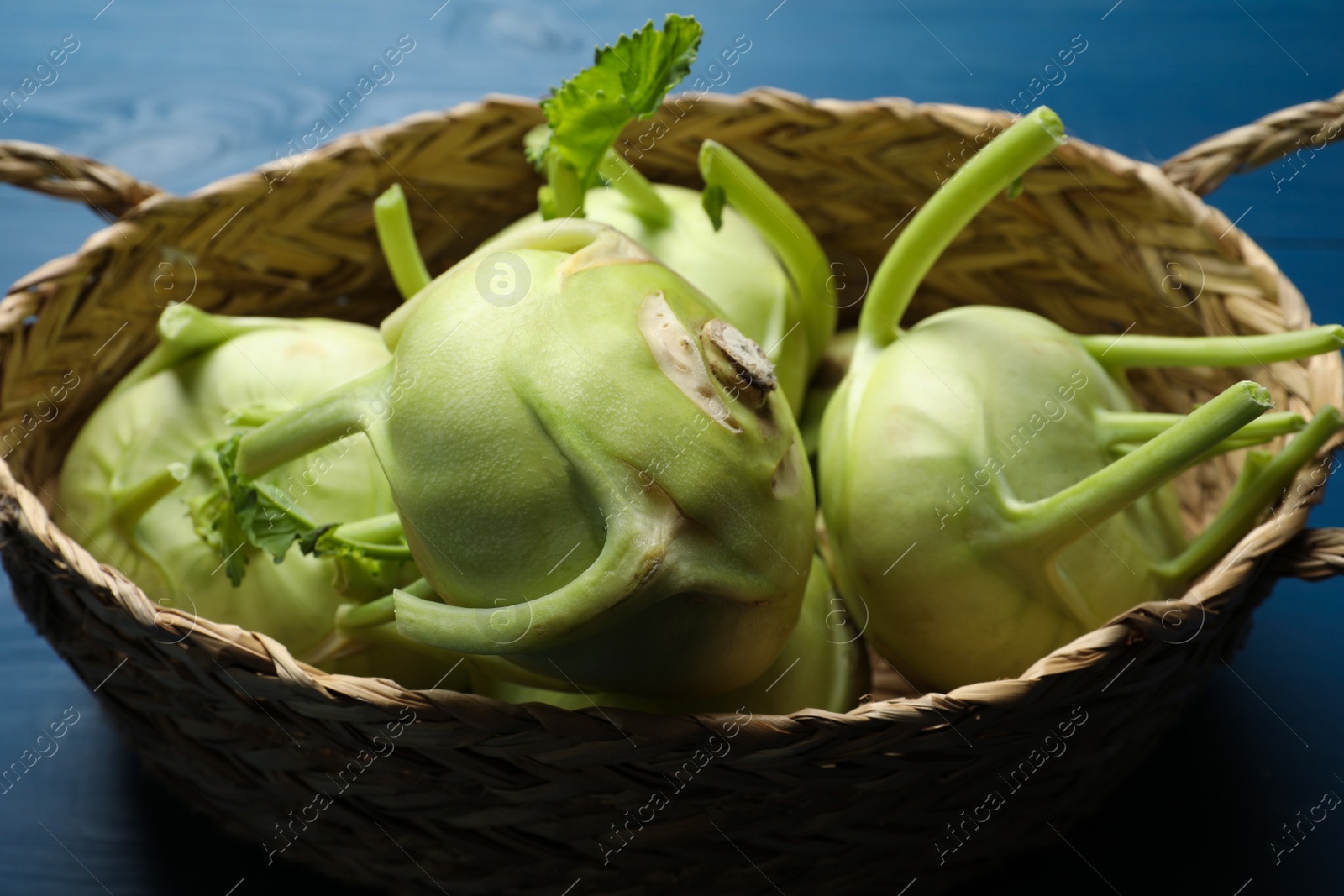 Photo of Whole ripe kohlrabi plants in wicker basket on blue table