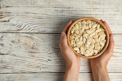 Photo of Young woman with bowl of raw pumpkin seeds at white wooden table, top view. Space for text