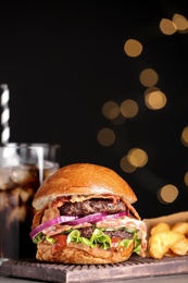 Board with burger and cola on table against blurred lights