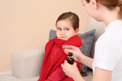 Photo of Mother giving cough syrup to her daughter from measuring cup on sofa indoors