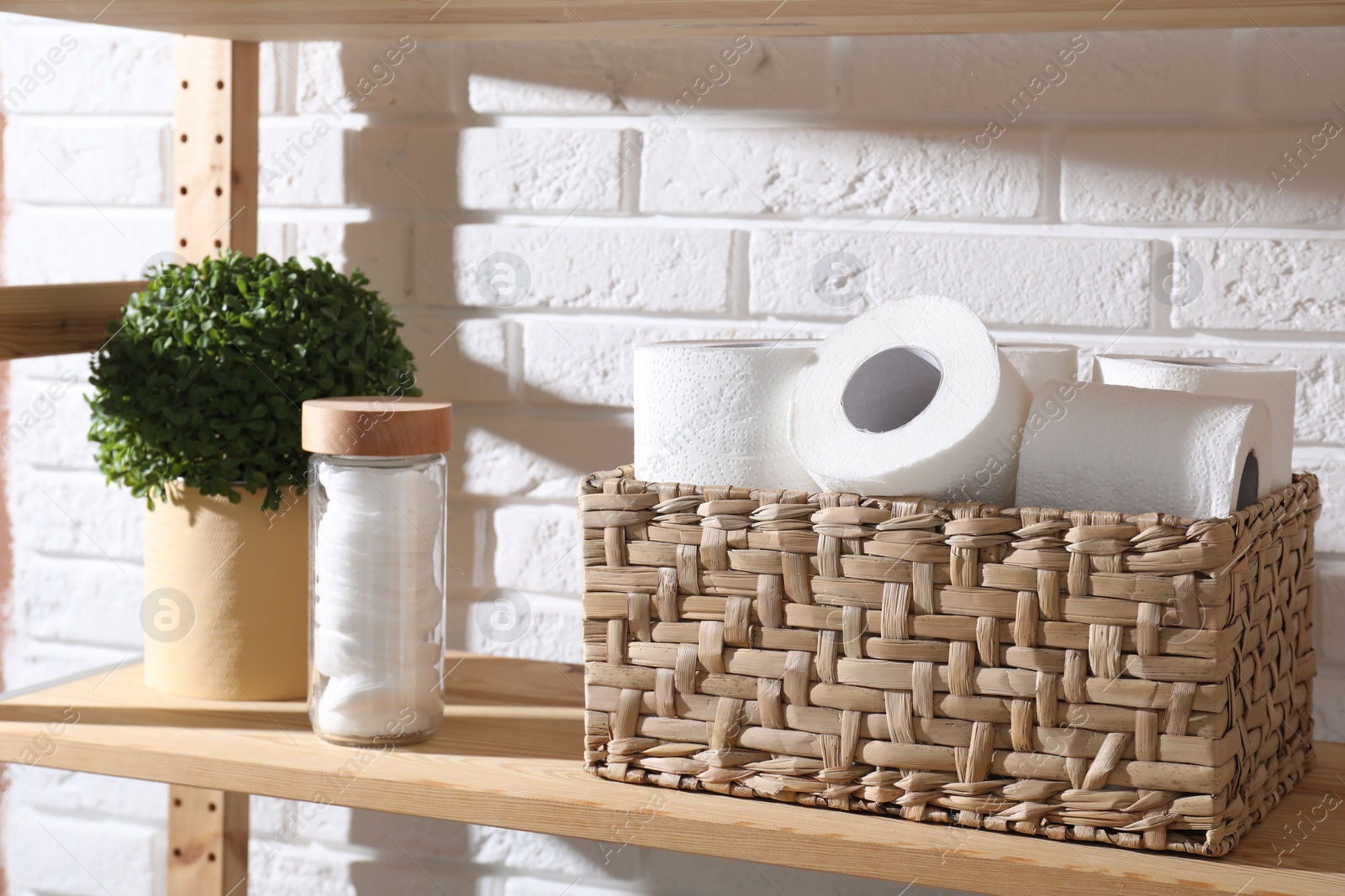 Photo of Toilet paper rolls in wicker basket, floral decor and cotton pads on wooden shelf near white brick wall