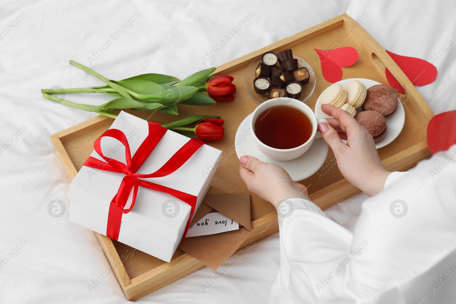 Photo of Tasty breakfast served in bed. Woman with tea, desserts, gift box and flowers at home, closeup