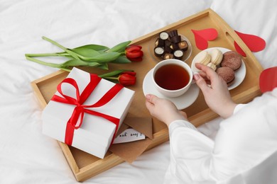 Photo of Tasty breakfast served in bed. Woman with tea, desserts, gift box and flowers at home, closeup