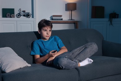 Photo of Little boy changing TV channels with remote control on sofa at home