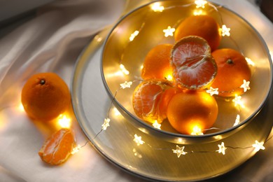 Photo of Fresh ripe tangerines and glowing fairy lights on white cloth, above view