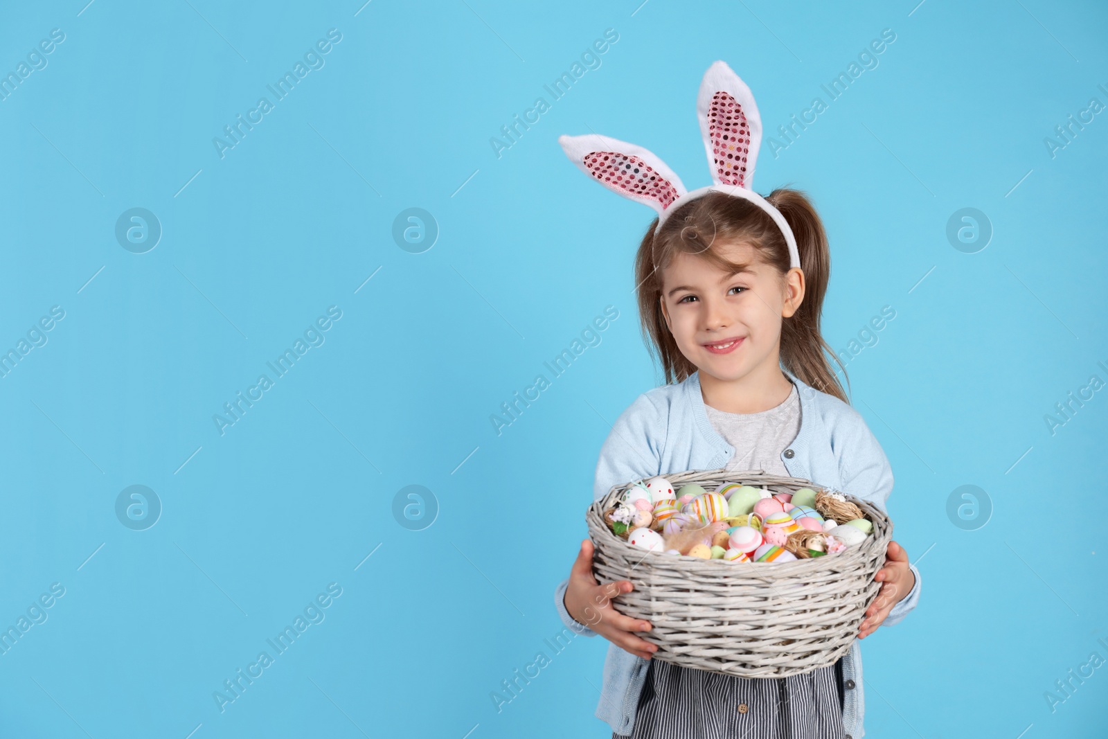 Photo of Happy little girl with bunny ears holding wicker basket full of Easter eggs on light blue background. Space for text