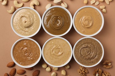 Many tasty nut butters in bowls and nuts on beige table, flat lay