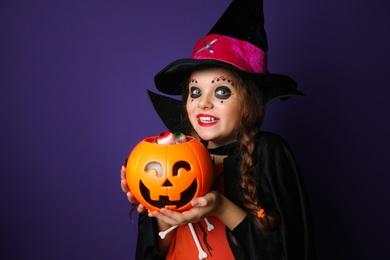 Photo of Cute little girl with pumpkin candy bucket wearing Halloween costume on purple background