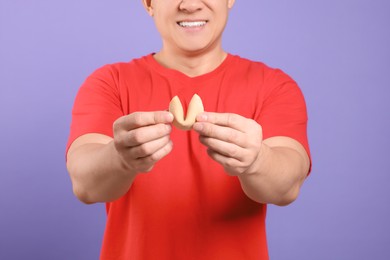 Happy man holding tasty fortune cookie with prediction on violet background, closeup