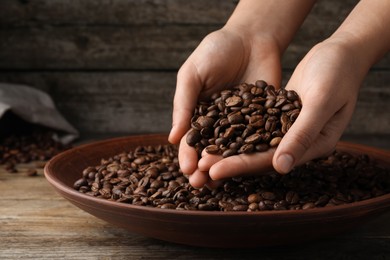 Photo of Woman taking pile of roasted coffee beans from bowl at wooden table, closeup