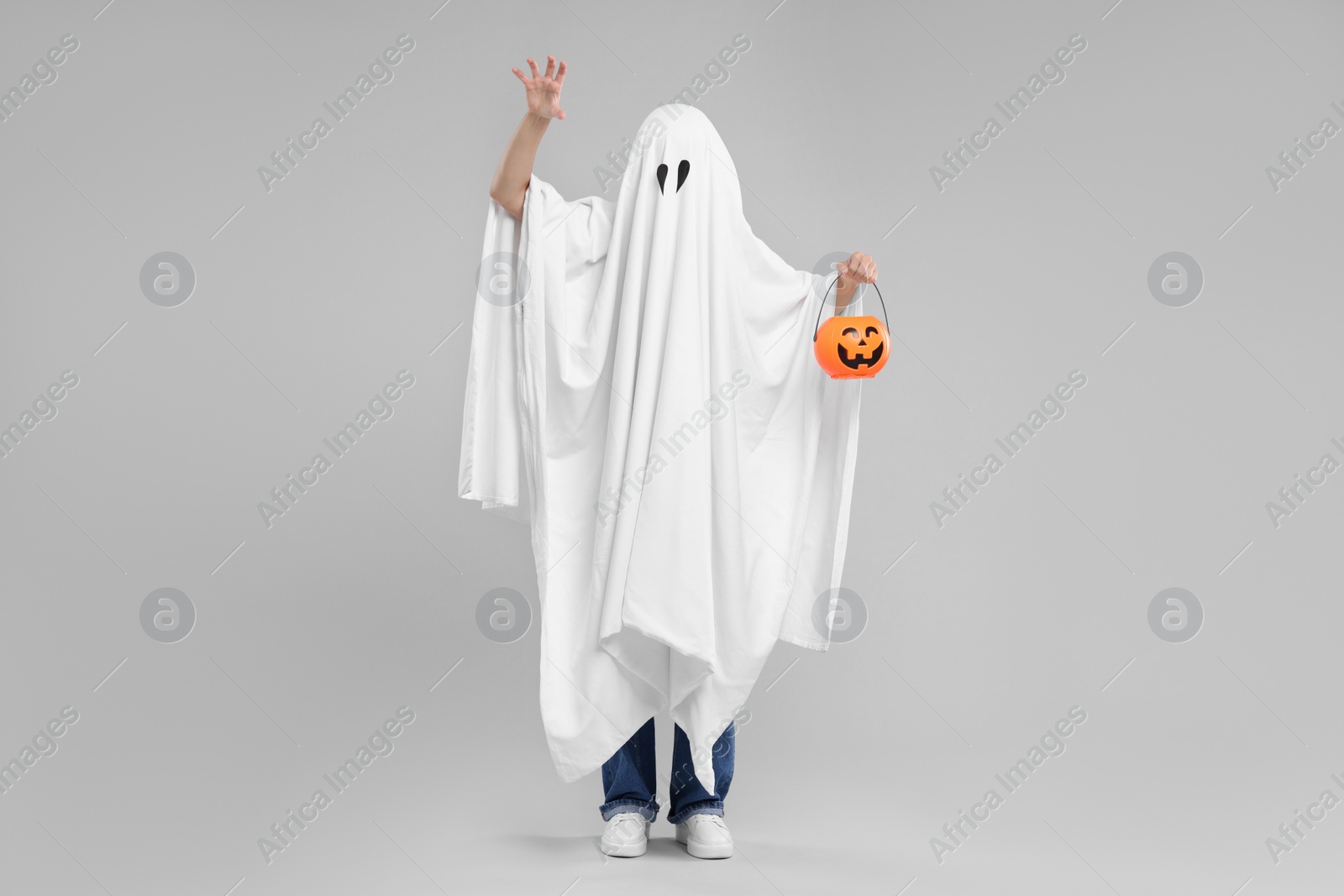 Photo of Woman in white ghost costume holding pumpkin bucket on light grey background. Halloween celebration